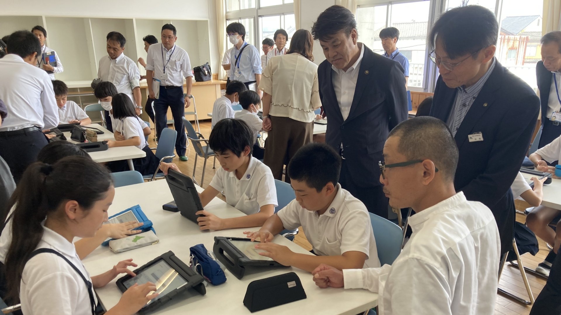 坂出市の有福哲二市長（左）、香川県商工労働部の寺嶋賢治部長（中央）、NextremerAIミナライ開発リーダー兼講師の行成晴（右）がAIを使って学ぶ児童を見守る様子